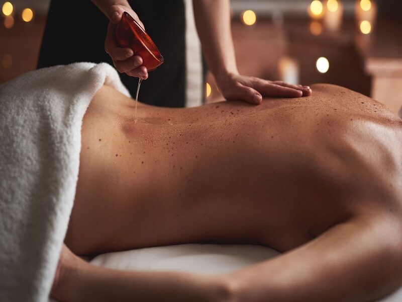 Concept of relaxation and body care. Cropped portrait of female massage therapist pouring essential oil out from bowl on back of young woman laying in spa salon