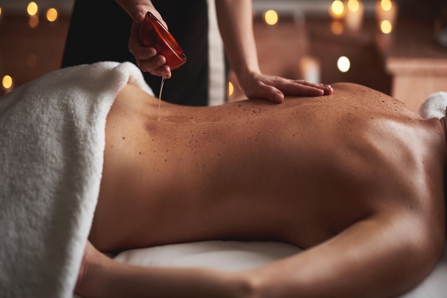 Concept of relaxation and body care. Cropped portrait of female massage therapist pouring essential oil out from bowl on back of young woman laying in spa salon